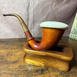 May include: A tobacco pipe with a polished amber-toned wooden bowl, a smooth off-white ceramic lid, and a curved translucent stem. The pipe rests securely within a carved, rectangular wooden stand featuring a natural grain finish.
