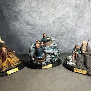 Puede incluir: Tres figuras detalladas de la serie El Hobbit. Una representa a Gollum y un hobbit, otra muestra personajes en un bote, y la tercera presenta a Gandalf con un hobbit. Cada figura está sobre una base ovalada negra.