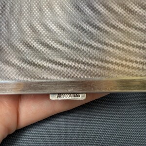 May include: Close-up of a silver metal object with a textured surface. The object has a rectangular shape with a small tag that reads "ALPACCA Hoka". The background is a dark gray fabric.