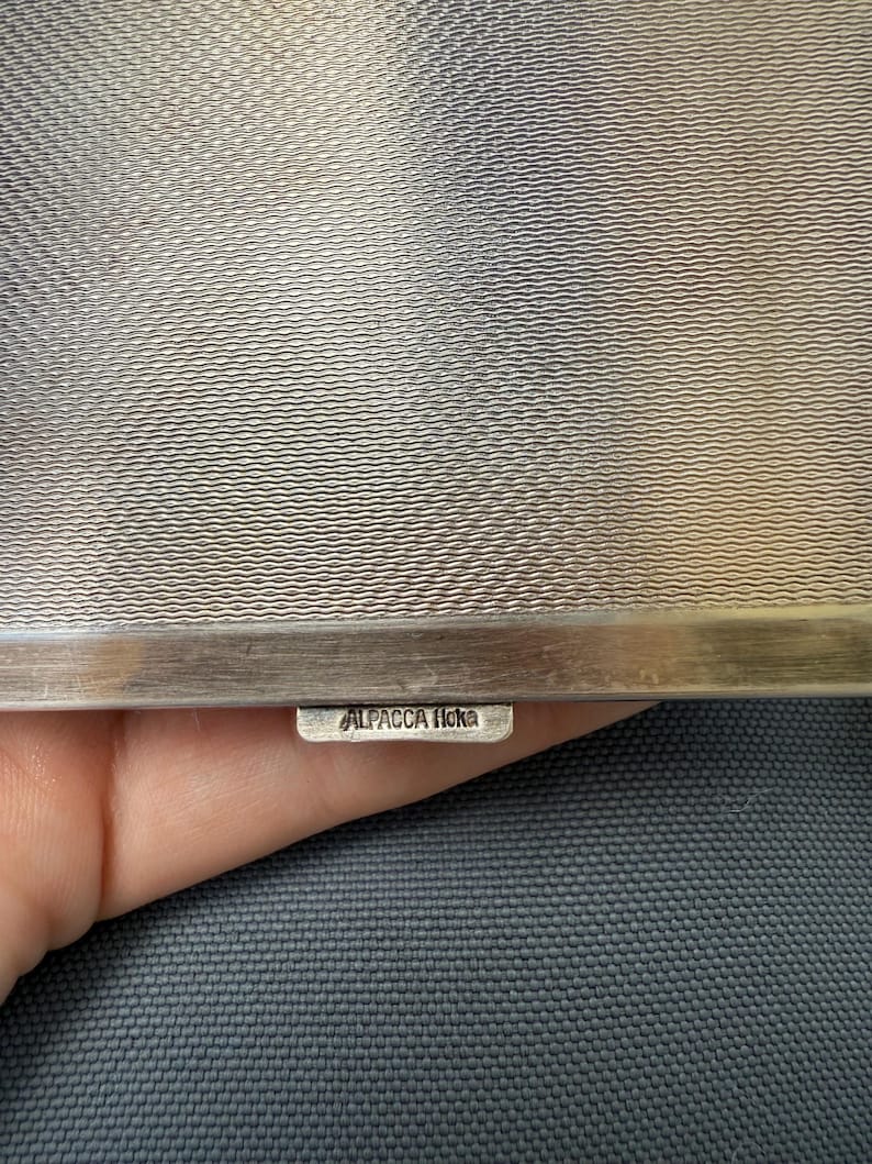 May include: Close-up of a silver metal object with a textured surface. The object has a rectangular shape with a small tag that reads "ALPACCA Hoka". The background is a dark gray fabric.