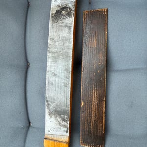 Puede incluir: Un cuero para navaja de afeitar recta vintage y una pieza de cuero separada. El cuero tiene un mango de madera y una superficie metálica, que muestra signos de desgaste. La pieza de cuero es de color marrón oscuro y rectangular. El cuero tiene el texto "GERMANY".