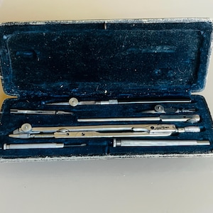Könnte beinhalten: Ein Vintage-Kompass-Set in einem schwarzen Samt-Etui. Das Set enthält einen Kompass, einen Teiler und einen Bleistift. Die Werkzeuge sind aus Silbermetall gefertigt und haben eine polierte Oberfläche.