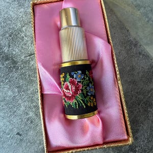 Puede incluir: Un frasco de perfume vintage con tapa plateada y una funda bordada en negro con un diseño floral en rojo, amarillo, azul y verde. El frasco se presenta en una caja con ribete dorado y forro de satén rosa.