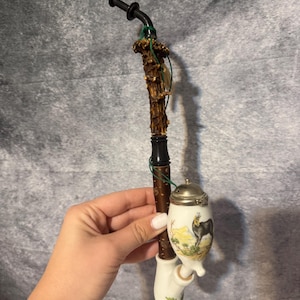 May include: A decorative tobacco pipe with a deer scene. The pipe features a ceramic bowl with a silver lid, a wooden stem, and a unique antler handle. The bowl is white with a painted landscape.
