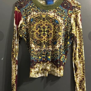 May include: A long-sleeved shirt with a colorful paisley pattern in yellow, blue, and burgundy. The shirt has a green ribbed crew neck and the brand name "KENZO JEANS" is visible on the collar.