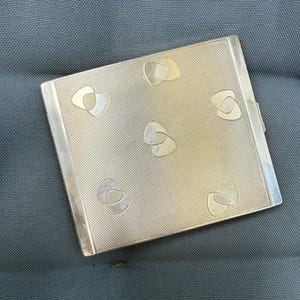 May include: A silver-toned compact with a textured surface and decorative abstract shapes. The rectangular compact has a hinged closure and is likely made of metal. The design features a repeating pattern of stylized, layered shapes.