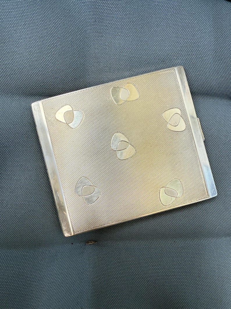 May include: A silver-toned compact with a textured surface and decorative abstract shapes. The rectangular compact has a hinged closure and is likely made of metal. The design features a repeating pattern of stylized, layered shapes.