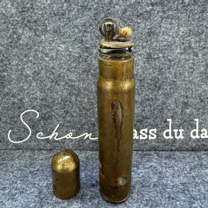 Könnte beinhalten: Ein Vintage-Feuerzeug aus Messing mit zylindrischem Körper und separater Kappe. Das Feuerzeug weist Gebrauchsspuren und Oxidation auf, was ihm ein gealtertes Aussehen verleiht. Die Oberseite hat einen Docht und einen kleinen, kreisförmigen Mechanismus. Die Kappe ist ein kleinerer, abgerundeter Zylinder.