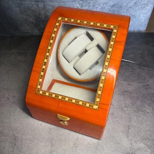 Puede incluir: Una caja giratoria para relojes de madera pulida color marrón anaranjado con tapa curva y ventana de cristal enmarcada por una incrustación de cuadros. El interior cuenta con un soporte giratorio circular y un compartimento de almacenamiento fijo, ambos forrados con tela gris claro, y un cierre de metal dorado con cerradura en la parte frontal.