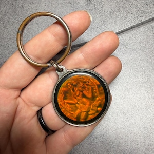 May include: A round keychain with a bronze-colored ring and a circular pendant. The pendant has a black border and an orange, swirling design. The keychain is held in a hand.