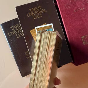 Pu&ograve; includere: Mazzo di Tarocchi Universal Dali con bordi dorati. Il mazzo &egrave; in una scatola marrone con il titolo in lettere dorate. La scatola &egrave; accanto a una scatola di velluto rosso con lo stesso titolo. Le carte sono visibili.