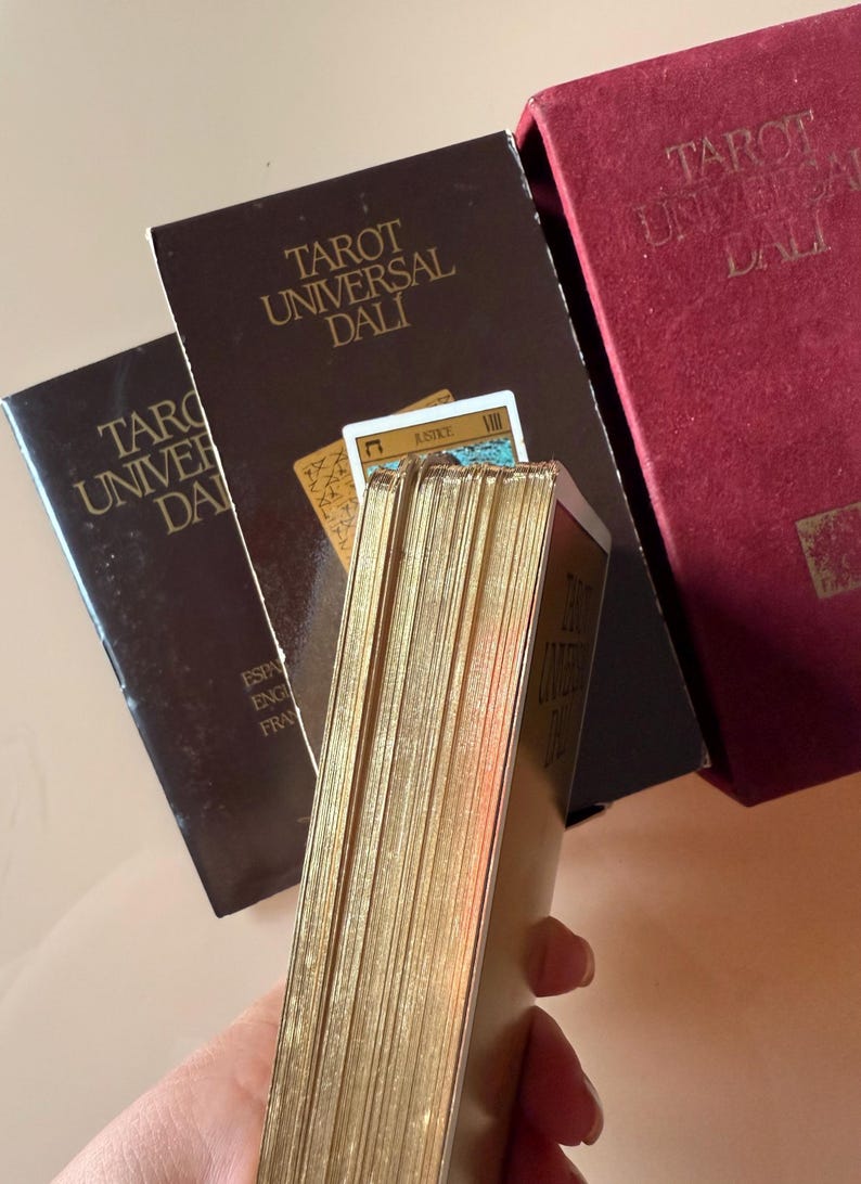 Pu&ograve; includere: Mazzo di Tarocchi Universal Dali con bordi dorati. Il mazzo &egrave; in una scatola marrone con il titolo in lettere dorate. La scatola &egrave; accanto a una scatola di velluto rosso con lo stesso titolo. Le carte sono visibili.