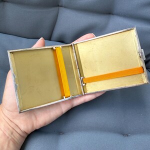May include: Open silver-toned cigarette case with a gold interior and two orange elastic bands. The case is rectangular and has a hinged lid. The interior is a light gold color, and the case is held open by a hand.