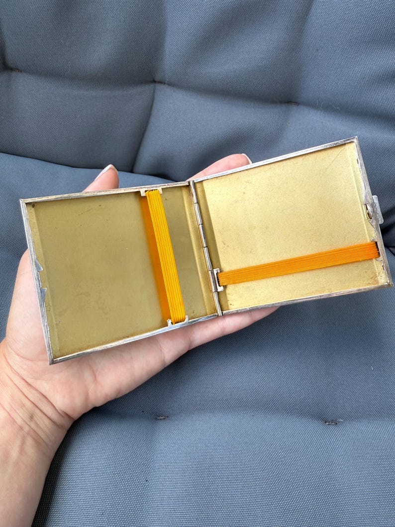May include: Open silver-toned cigarette case with a gold interior and two orange elastic bands. The case is rectangular and has a hinged lid. The interior is a light gold color, and the case is held open by a hand.