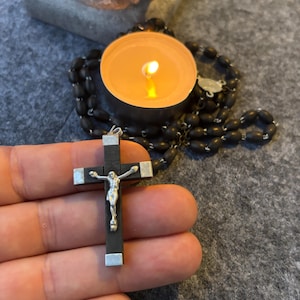May include: A black wooden crucifix with silver detailing, depicting a silver figure of Jesus. A black beaded rosary and a lit tea light candle are also present. The items are on a grey surface.