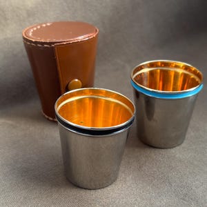 Könnte beinhalten: Zwei silberfarbene Schnapsgläser mit goldfarbenem Innenraum und einem braunen Lederetui. Ein Glas hat ein schwarzes Band, das andere ein blaues Band. Das Etui ist zylindrisch mit einem Druckknopfverschluss. Die Gläser sind ca. 5 cm hoch.