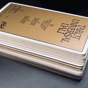 Pu&ograve; includere: Un mazzo di carte dei Tarocchi intitolato "Tarot Universal Dali" con una carta superiore color oro. Il mazzo ha bordi bianchi ed &egrave; impilato su una superficie scura. La carta superiore presenta testo in inglese e spagnolo.