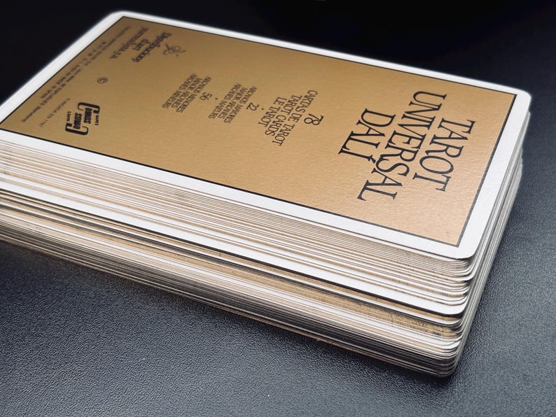 Pu&ograve; includere: Un mazzo di carte dei Tarocchi intitolato "Tarot Universal Dali" con una carta superiore color oro. Il mazzo ha bordi bianchi ed &egrave; impilato su una superficie scura. La carta superiore presenta testo in inglese e spagnolo.