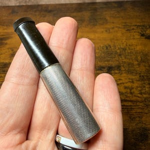 May include: A cigarette holder featuring a black mouthpiece and a cylindrical silver-toned body with a fine, cross-hatched, engine-turned texture. The metallic section has a polished finish, while the mouthpiece is smooth and dark.