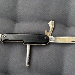 Pode incluir: Uma canivete multi-ferramentas vintage com cabo preto e várias ferramentas dobráveis. As ferramentas incluem um abridor de garrafas, um saca-rolhas e uma pequena lâmina. A pega tem texto que é difícil de ler. A faca está numa superfície cinzenta.