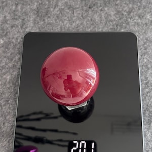 Op de afbeelding: Een glanzend, rond, dieprood object ligt op een zwarte digitale weegschaal, met een gewicht van 201. De weegschaal heeft een digitale display en de merknaam "Yilai". Het object reflecteert de omgeving.