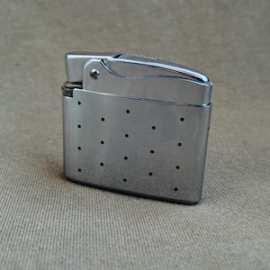 May include: A vintage chrome lighter with a rectangular body and a hinged top. The lighter features a series of small, evenly spaced dots on the front and a textured surface on the top. The lighter is a silver color.