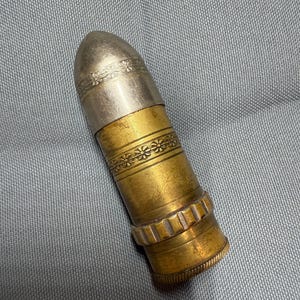 May include: A vintage bullet-shaped container with a silver top and brass body. The silver top features floral engravings, while the brass section has decorative bands. The base has a ridged design.