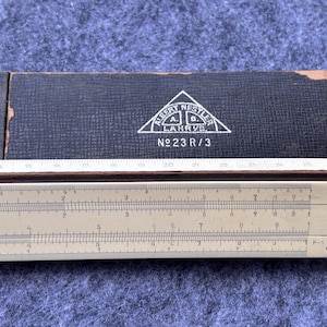 May include: A vintage slide rule with a cream-coloured body and black markings, partially extended from its dark blue case. The case has a white triangle logo with text and the number "No 23 R/3".