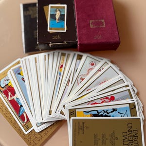 Pu&ograve; includere: Un mazzo di carte Tarot Universal Dali con una scatola bordeaux e due scatole marroni. Le carte sono a ventaglio, mostrando opere d'arte surrealiste colorate. Il testo "TAROT UNIVERSAL DALI" &egrave; visibile su una delle carte.
