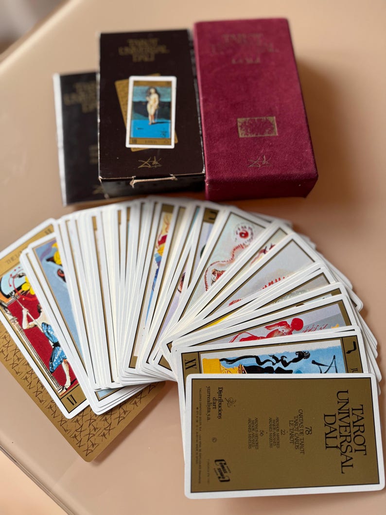 Pu&ograve; includere: Un mazzo di carte Tarot Universal Dali con una scatola bordeaux e due scatole marroni. Le carte sono a ventaglio, mostrando opere d'arte surrealiste colorate. Il testo "TAROT UNIVERSAL DALI" &egrave; visibile su una delle carte.