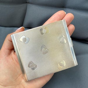 May include: A silver compact with a textured surface and decorative heart-shaped designs. The compact is rectangular and has a clasp on the side. The design features a repeating pattern of interlocking shapes in a lighter silver tone.