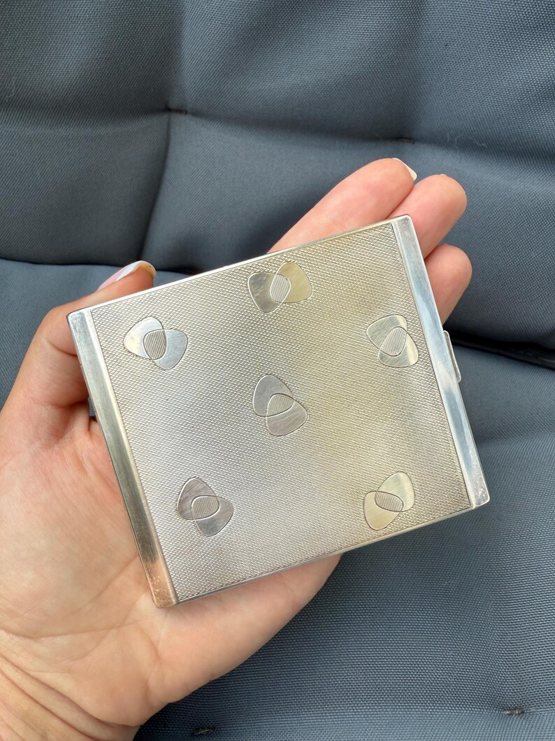 May include: A silver compact with a textured surface and decorative heart-shaped designs. The compact is rectangular and has a clasp on the side. The design features a repeating pattern of interlocking shapes in a lighter silver tone.
