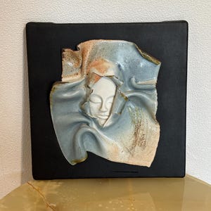 Può includere: Un'opera d'arte murale in ceramica raffigurante un volto sereno che emerge da forme drappeggiate e fluide. La scultura presenta un volto bianco, con accenti blu, marroni e marroni, su uno sfondo quadrato nero. L'opera d'arte è esposta su un tavolo.