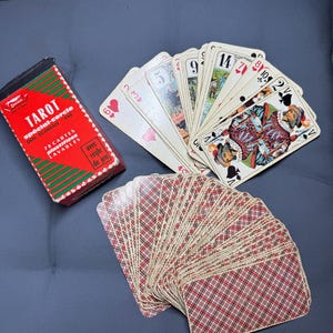 May include: A deck of 78 tarot cards with a red and green box. The cards are fanned out, showing various suits and illustrations. The card backs have a red and brown plaid pattern. The box reads "TAROT" and "78 cartes plastifiées lavables".