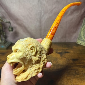 May include: A tobacco pipe featuring a carved cream-colored monkey head bowl with a textured surface and an open mouth. The pipe has a long, curved stem made of translucent orange and yellow marbled material with a smooth, glossy finish.