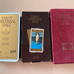 Pu&ograve; includere: Tre scatole di carte dei tarocchi e un mazzo di carte. Le scatole sono etichettate "Tarot Universal Dali" in lettere dorate e bianche. Il mazzo di carte presenta una donna in piedi davanti a uno sfondo blu. Le scatole sono bordeaux, marroni e dorate.