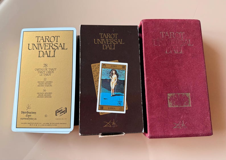 Pu&ograve; includere: Tre scatole di carte dei tarocchi e un mazzo di carte. Le scatole sono etichettate "Tarot Universal Dali" in lettere dorate e bianche. Il mazzo di carte presenta una donna in piedi davanti a uno sfondo blu. Le scatole sono bordeaux, marroni e dorate.