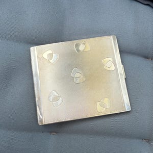 May include: A rectangular silver-toned cigarette case with a textured surface and six decorative, abstract shapes. The case has a hinged lid and a polished silver border. The case is resting on a gray surface.