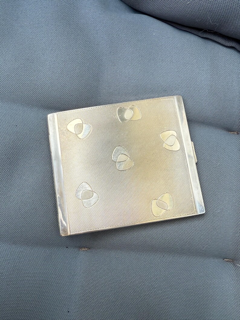 May include: A rectangular silver-toned cigarette case with a textured surface and six decorative, abstract shapes. The case has a hinged lid and a polished silver border. The case is resting on a gray surface.