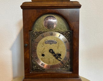 Può includere: Un orologio da camino in legno con finitura marrone scuro, dotato di un quadrante dorato con numeri romani e lancette nere. L'orologio riporta la scritta Tempus Fugit in alto e Warming al centro. Include una maniglia in ottone sulla parte superiore.