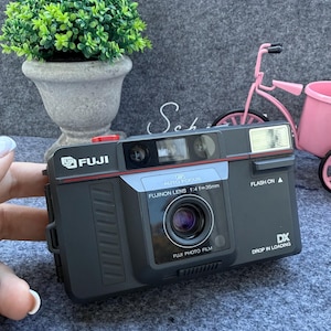 Puede incluir: Una cámara Fuji negra con una línea de acento roja, detalles plateados y el texto "FUJI" y "FUJINON LENS". La cámara tiene una lente, flash y el texto "DX DROP IN LOADING". Una pequeña planta verde en una maceta gris está en el fondo.
