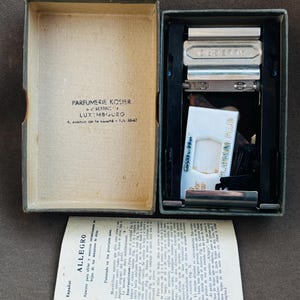 Peut inclure: Un étui de rasoir vintage ouvert, avec un rasoir et une lame Gillette Platinum Plus. L'étui est gris foncé avec une boîte en carton à gauche. La boîte contient du texte en français. Une notice pliée est en espagnol.