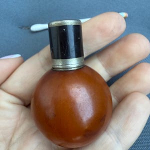 以下が含まれることがあります： 黒いキャップと銀色のトリムが付いた、小さくて丸い琥珀色のボトル。ボトルは青い背景に対して手で持たれています。青とオレンジの先端が付いた小さな白いツールが背景にあります。