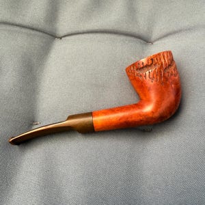 Peut inclure: Une pipe à tabac brun rougeâtre avec une tige incurvée et un foyer sculpté. La pipe a un embout buccal brun foncé et une bague couleur laiton reliant la tige au foyer. Le foyer présente un motif texturé et sculpté.