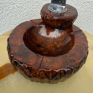 Può includere: Un set di posacenere vintage con un accendino abbinato. Il posacenere è di colore marrone scuro, di forma circolare, con un'area incassata per le ceneri. L'accendino è cromato e poggia su una base marrone più piccola abbinata. Il set ha una finitura lucida.