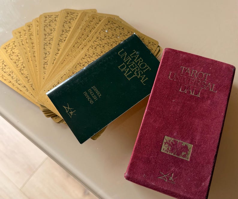 Pu&ograve; includere: Un mazzo di carte dei tarocchi con dorsi dorati, a ventaglio accanto a due libri. Un libro &egrave; verde scuro con il testo "Tarot Universal Dali" e "Espa&ntilde;ol English Fran&ccedil;ais". L'altro libro &egrave; bordeaux con lo stesso titolo.