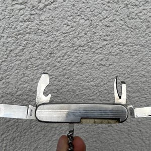 May include: A silver multi-tool with several open blades and tools, including a knife, bottle opener, and corkscrew. The tool has a textured handle and is partially open, displaying its various functions. The image is taken against a textured gray background.