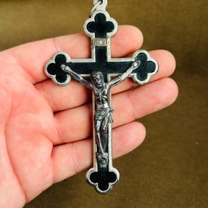 Può includere: Ciondolo a forma di crocifisso argentato con croce nera e figura dettagliata di Gesù. La croce presenta estremità decorative a trifoglio e un anello per appenderlo. La figura è scolpita in modo intricato, raffigurante la crocifissione.