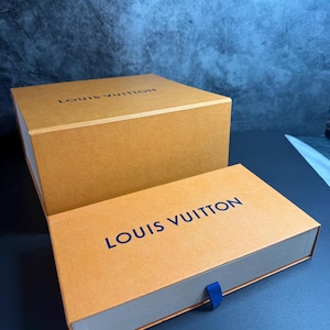May include: Two rectangular Louis Vuitton boxes in a signature tan color with dark blue lettering. The larger box is positioned behind a smaller, flatter box that features a small blue fabric pull tab on the front edge for opening the drawer.