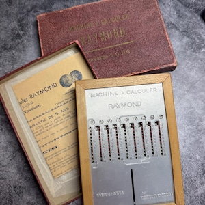 Peut inclure: Calculatrice mécanique Raymond vintage avec un cadre en bois et une face en métal. La calculatrice comporte des cadrans numériques et un code couleur rouge et argent. La boîte et le manuel d'instructions sont également visibles.